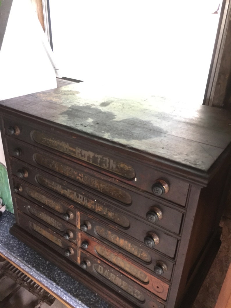 May include: A dark wooden antique drawer cabinet with eight drawers. Each drawer has a glass front and a metal knob. The drawers are labeled with faded text, including "Cotton", "Dark", and "Arc".