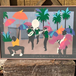 Puede incluir: Una impresi&oacute;n enmarcada de una ilustraci&oacute;n colorida que representa una escena callejera de Nueva Orleans con una banda de m&uacute;sica, palmeras y personas con trajes coloridos. El texto "NEW ORLEANS" est&aacute; impreso en la parte inferior de la impresi&oacute;n.