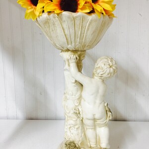 May include: A cream-colored cherub statue holding a decorative bowl filled with yellow sunflowers. The statue has a detailed design and is made of a stone-like material. The sunflowers have dark brown centers.