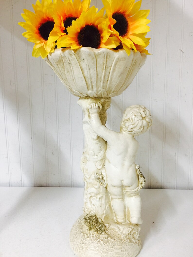 May include: A cream-colored cherub statue holding a decorative bowl filled with yellow sunflowers. The statue has a detailed design and is made of a stone-like material. The sunflowers have dark brown centers.