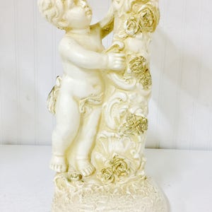 May include: A cream-colored cherub figurine, holding onto a decorative column adorned with roses. The base has a textured, aged appearance. The cherub is sculpted in a classical style, with detailed facial features and curly hair. A decorative accent piece.