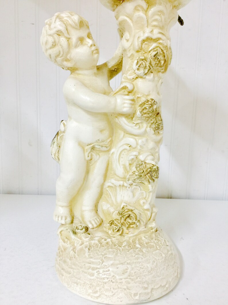 May include: A cream-colored cherub figurine, holding onto a decorative column adorned with roses. The base has a textured, aged appearance. The cherub is sculpted in a classical style, with detailed facial features and curly hair. A decorative accent piece.