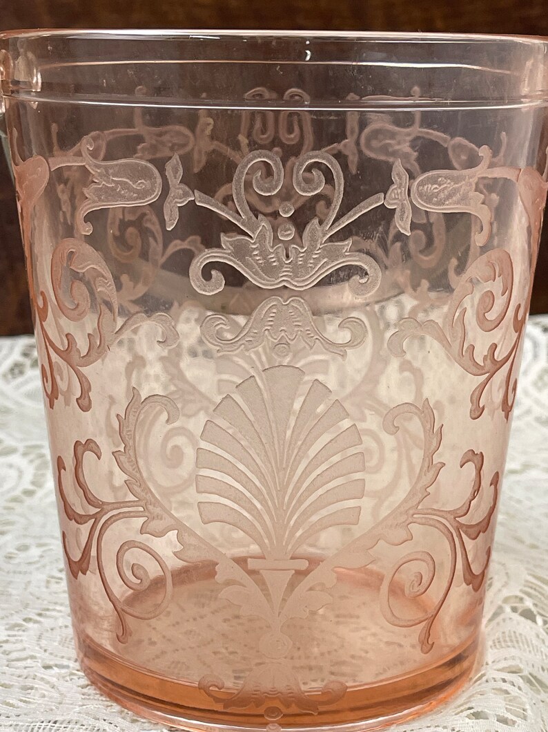 May include: A pink glass ice bucket with an etched floral design. The design features a large shell in the centre and smaller floral motifs around the edges.