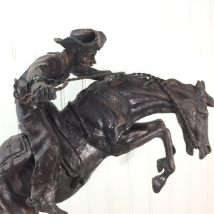 May include: A bronze sculpture of a cowboy riding a bucking horse. The cowboy is wearing a hat and is holding onto the reins. The horse is rearing up on its hind legs.