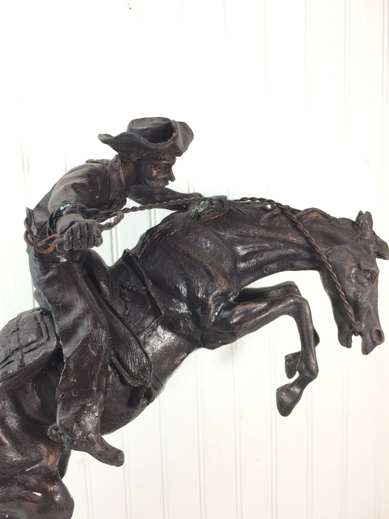 May include: A bronze sculpture of a cowboy riding a bucking horse. The cowboy is wearing a hat and is holding onto the reins. The horse is rearing up on its hind legs.