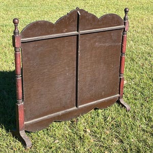 May include: A dark brown wooden screen with a decorative top and red accents on the legs. The screen has a cross-shaped design and stands on two feet. The screen is outdoors on a grassy surface.