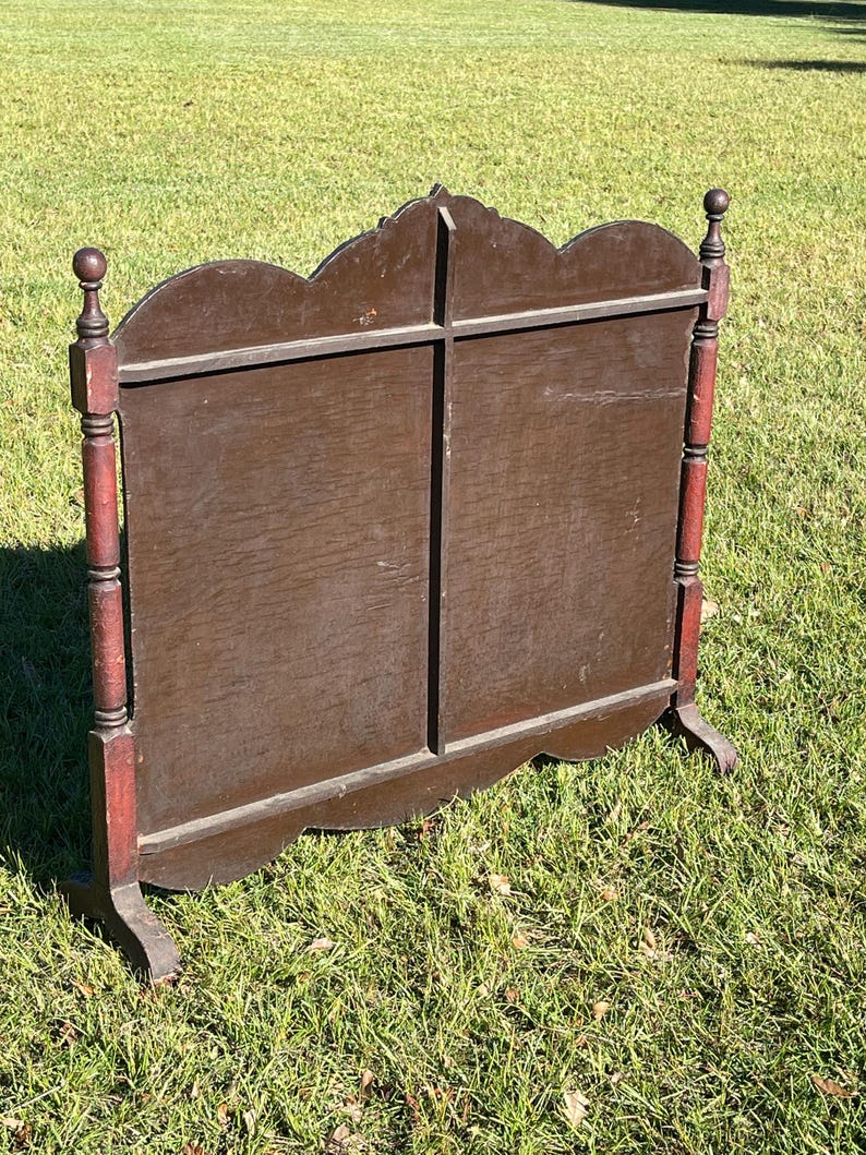May include: A dark brown wooden screen with a decorative top and red accents on the legs. The screen has a cross-shaped design and stands on two feet. The screen is outdoors on a grassy surface.