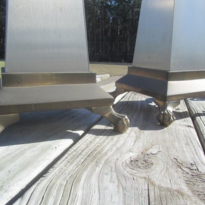 May include: Two silver and brass table lamps with a square base and four feet. The lamps are on a wooden surface.