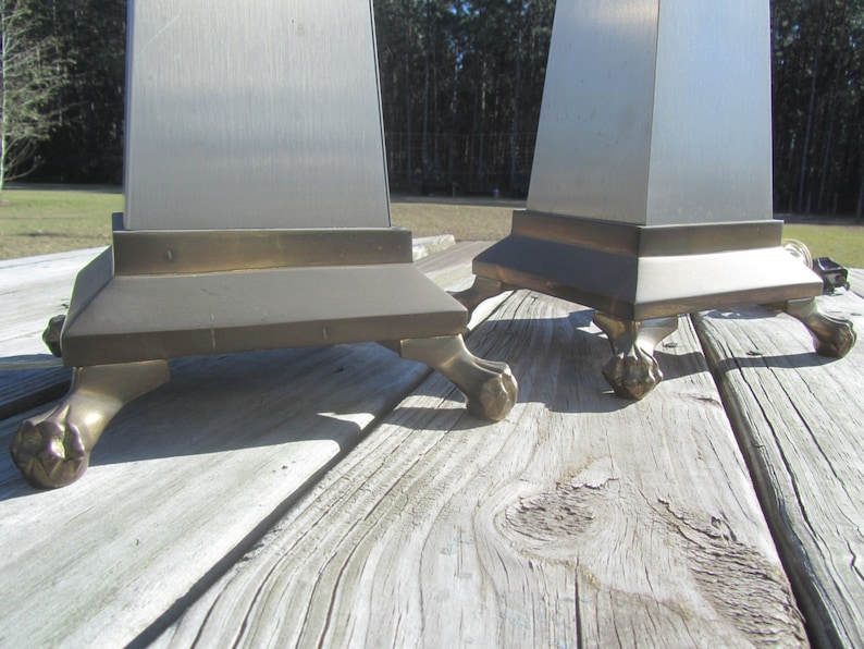 May include: Two silver and brass table lamps with a square base and four feet. The lamps are on a wooden surface.