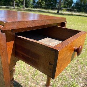 May include: A wooden antique drop-leaf table with a drawer open, revealing its interior. The table is a warm brown color, with a round wooden knob on the drawer. The table legs are turned and have a decorative design. The table is outdoors.