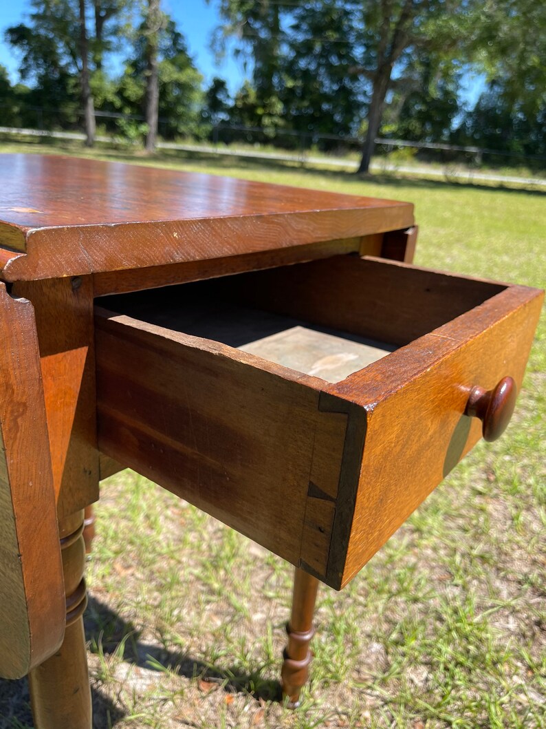 May include: A wooden antique drop-leaf table with a drawer open, revealing its interior. The table is a warm brown color, with a round wooden knob on the drawer. The table legs are turned and have a decorative design. The table is outdoors.