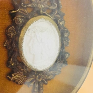 May include: An ornate gold-toned frame with a white cameo inside. The cameo features a raised design of a person's face.