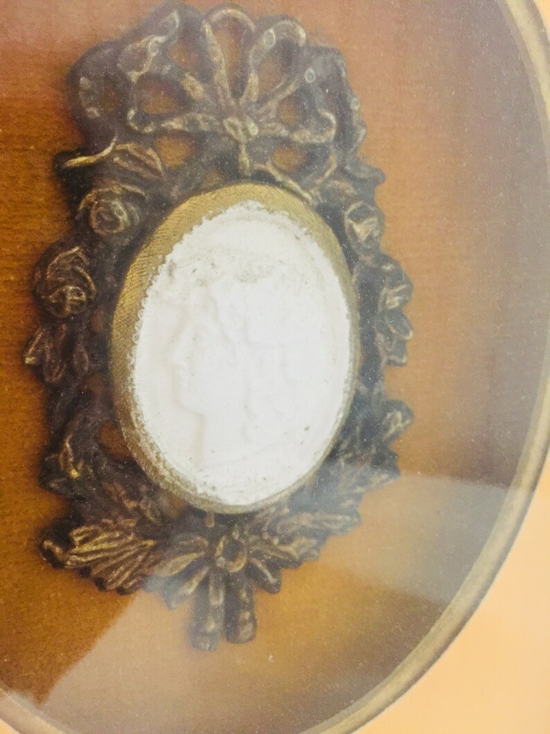 May include: An ornate gold-toned frame with a white cameo inside. The cameo features a raised design of a person's face.