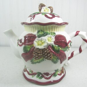 May include: White ceramic teapot with a red and white candy cane handle. The teapot is decorated with a winter scene of pine cones, holly, and red bows.