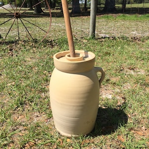 May include: A vintage cream-colored ceramic butter churn with a wooden paddle. The churn has a handle and a lid. The churn is sitting on a bed of green grass.