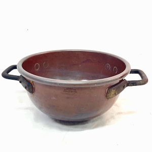 May include: A round copper basin with a smooth, dark metallic finish and two dark, weathered iron handles attached to the sides with rivets. The interior rim features visible rivet marks, and a small stamped maker mark is present on the front.