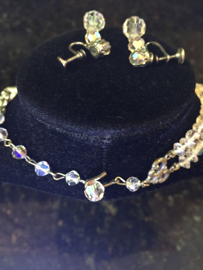 May include: A crystal necklace and earrings set with a silver chain and clear crystal beads. The necklace has a clasp closure. The earrings are clip-on style.