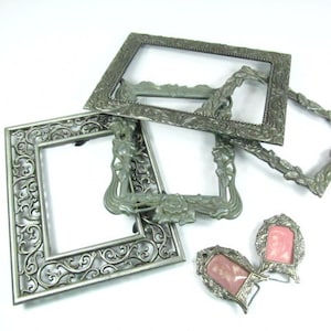 May include: Four silver-toned picture frames with ornate designs. Two of the frames are smaller and have a pink cameo inside. The frames are arranged on a white background.