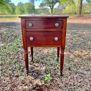 May include: A two-drawer wooden side table with turned legs. The table has a dark brown finish and silver drawer pulls. The table is sitting on a bed of brown leaves.