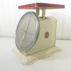 Op de afbeelding: Vintage witte keukenweegschaal met een rode bovenkant en een zilveren wijzerplaat. De weegschaal heeft een "Sunbeam"-logo aan de zijkant.