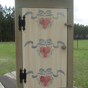 May include: A small, green painted wooden cabinet with a hinged door. The door is decorated with a stencil of three red hearts with blue ribbons and green leaves. The cabinet has black metal hinges and a knob.