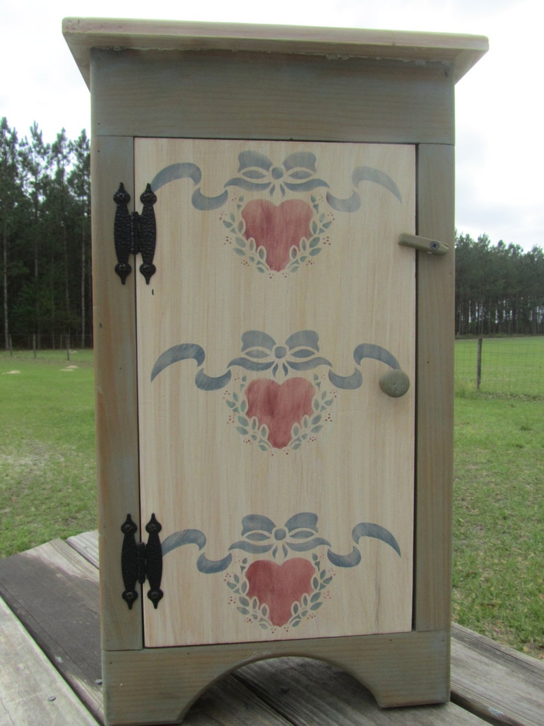May include: A small, green painted wooden cabinet with a hinged door. The door is decorated with a stencil of three red hearts with blue ribbons and green leaves. The cabinet has black metal hinges and a knob.