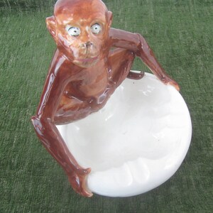 May include: A brown ceramic monkey figurine holding a white ceramic ashtray. The monkey has large eyes and a detailed face.