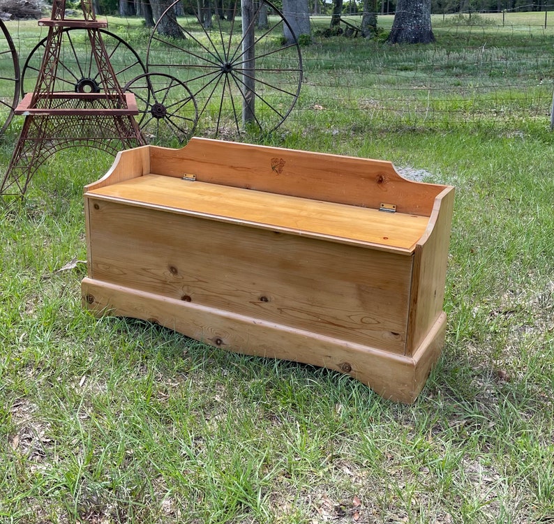 K&ouml;nnte beinhalten: Eine Holzaufbewahrungsbank mit einem Klappdeckel. Die Bank ist aus hellbraunem Holz gefertigt und hat ein schlichtes, rustikales Design.