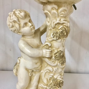 May include: A cream-colored cherub statue holding a decorative floral element. The statue features a detailed design with gold accents on the floral elements and the cherub's clothing. The cherub is standing next to a column with floral and scrollwork details.
