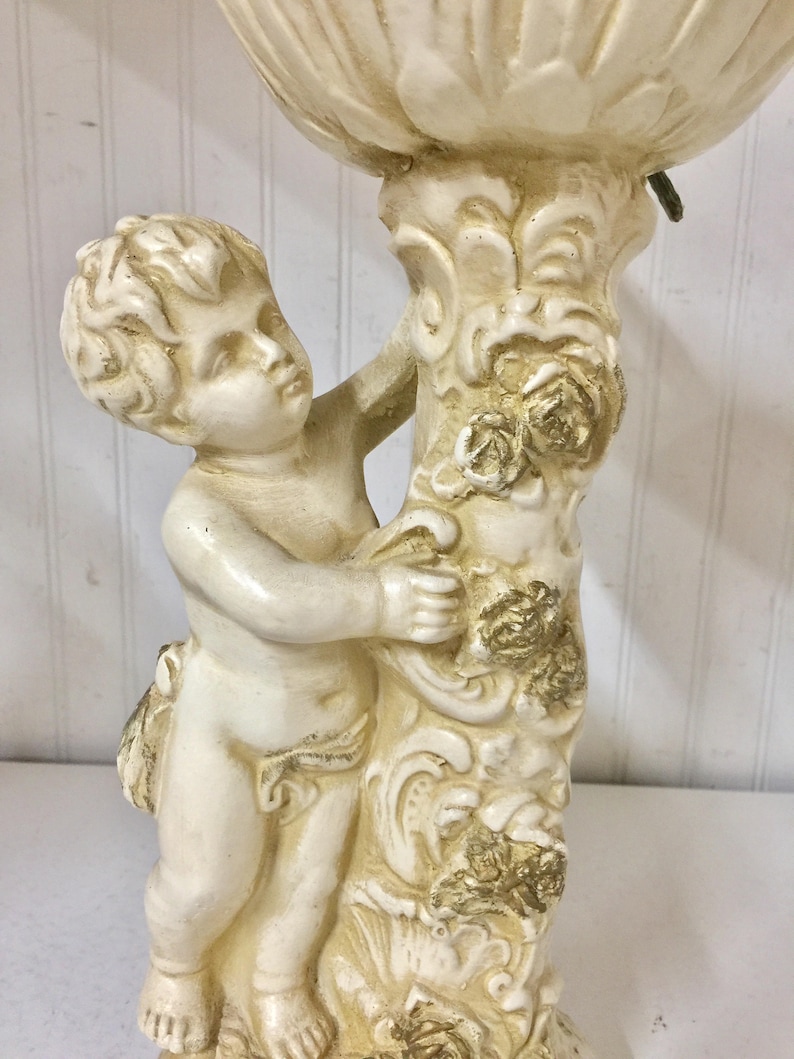 May include: A cream-colored cherub statue holding a decorative floral element. The statue features a detailed design with gold accents on the floral elements and the cherub's clothing. The cherub is standing next to a column with floral and scrollwork details.