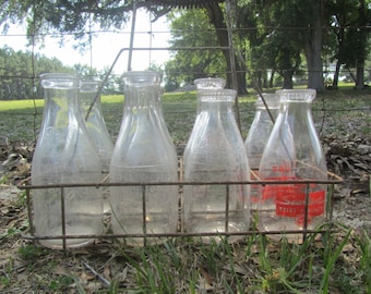 May include: A metal milk bottle carrier holding six clear glass milk bottles. One bottle has a red label with white text. The bottles are in a rusty metal carrier, set against a backdrop of green grass and trees.