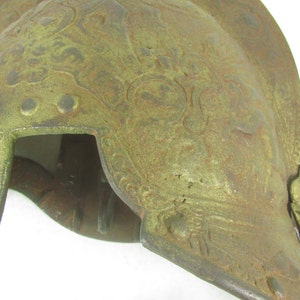 May include: A green metal helmet with intricate embossed designs. The helmet has a rounded top and a pointed front.