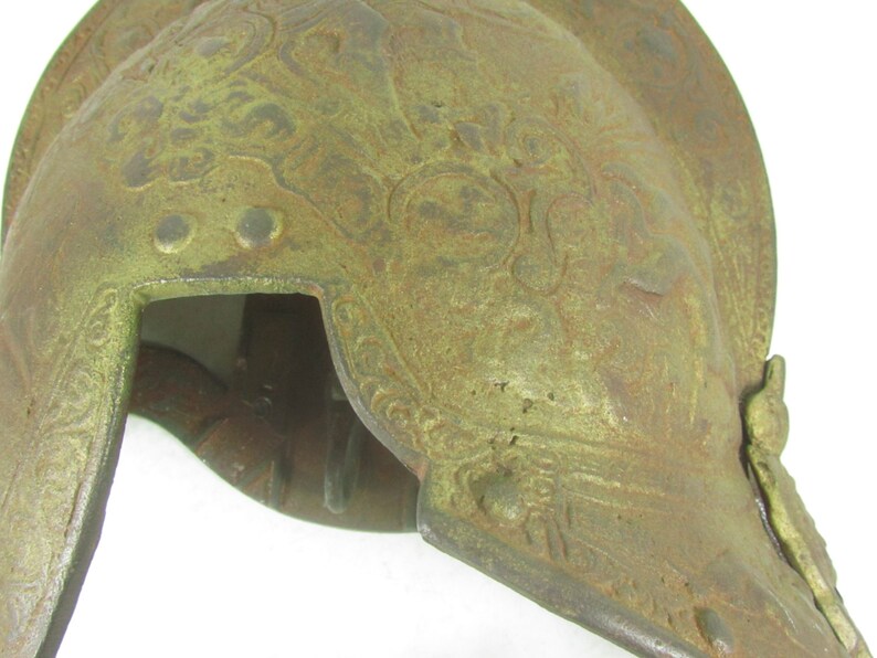 May include: A green metal helmet with intricate embossed designs. The helmet has a rounded top and a pointed front.