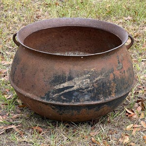 May include: A large, antique cast iron cauldron with a rounded body and a wide, open top. The cauldron has two small handles on either side and shows signs of rust and wear, with a dark, mottled patina. It sits on a bed of green grass.