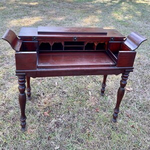 May include: A dark wood writing desk with a drop-down front and multiple drawers. The desk has turned legs and is sitting on a green grassy surface.