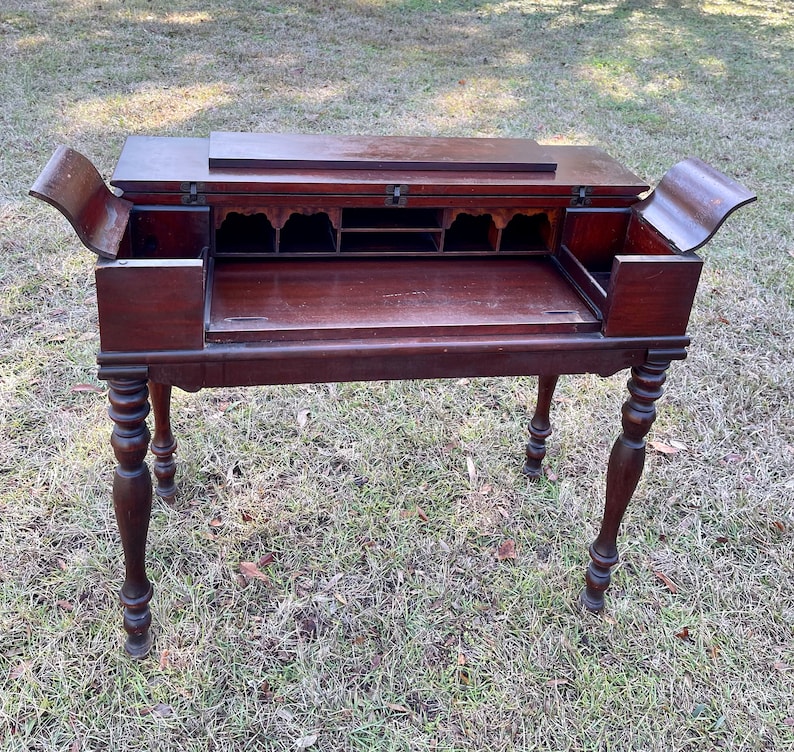 May include: A dark wood writing desk with a drop-down front and multiple drawers. The desk has turned legs and is sitting on a green grassy surface.