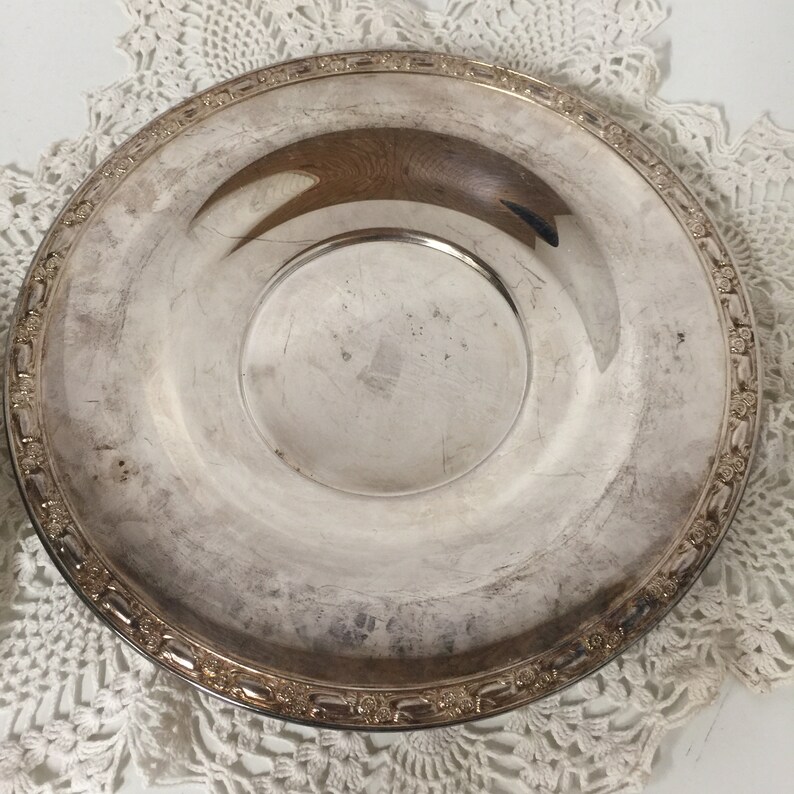 Puede incluir: Una bandeja de servir plateada con un borde decorativo. La bandeja tiene un centro ligeramente elevado y est&aacute; hecha de un metal brillante.