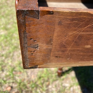 May include: Close-up of a wooden drawer corner, showcasing dovetail joints. The wood has a rich, dark brown color with visible grain and minor imperfections. The drawer is open, revealing the interior and construction details.