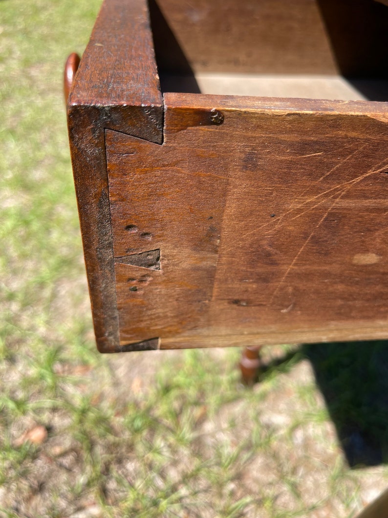 May include: Close-up of a wooden drawer corner, showcasing dovetail joints. The wood has a rich, dark brown color with visible grain and minor imperfections. The drawer is open, revealing the interior and construction details.