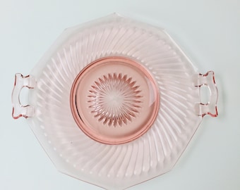 May include: A pink glass serving tray with an octagonal shape and two handles. The tray features a central, raised circular area with a starburst design and spiral grooves radiating outwards. The overall color is a soft, translucent pink.