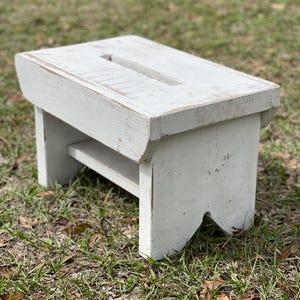 May include: A white wooden step stool with a distressed finish. The stool has a small shelf and a cutout in the top.