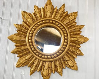 May include: Gold sunburst mirror with a round, beveled glass mirror in the center. The mirror is surrounded by a decorative, gold-painted frame with a sunburst design.