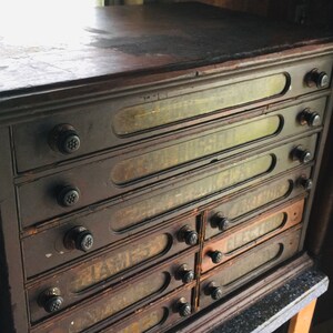 May include: A dark brown wooden cabinet with eight drawers. Each drawer has a glass front with faded labels. The cabinet has black knobs on each drawer.
