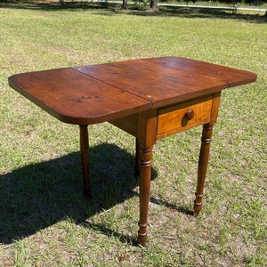 May include: A small, antique wooden drop-leaf table with a warm brown finish. The table features a single drawer with a round wooden knob and turned legs. The tabletop measures approximately 30 inches long when open.