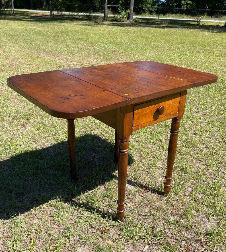 May include: A small, antique wooden drop-leaf table with a warm brown finish. The table features a single drawer with a round wooden knob and turned legs. The tabletop measures approximately 30 inches long when open.