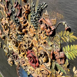 May include: A detailed, carved wooden floral relief. The artwork features a variety of flowers, including roses and sunflowers, in shades of brown, red, and yellow. The flowers are arranged in a blue vase against a dark background.