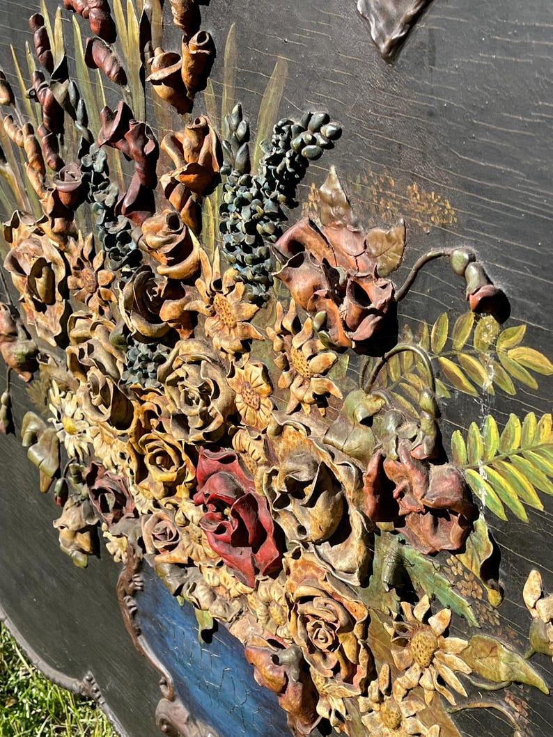 May include: A detailed, carved wooden floral relief. The artwork features a variety of flowers, including roses and sunflowers, in shades of brown, red, and yellow. The flowers are arranged in a blue vase against a dark background.