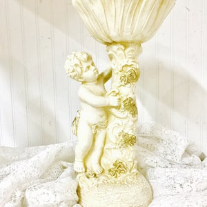 May include: A cream-colored cherub statue holding a large, petal-shaped bowl. The statue has gold accents on the column and base. The cherub is detailed with curly hair and delicate features. The statue is on a white lace cloth.
