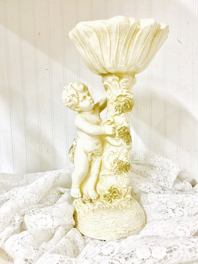 May include: A cream-colored cherub statue holding a large, petal-shaped bowl. The statue has gold accents on the column and base. The cherub is detailed with curly hair and delicate features. The statue is on a white lace cloth.