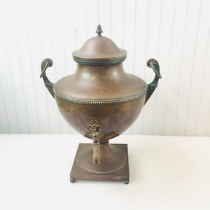 May include: Antique copper tea kettle with ornate handles and a spout. The kettle has a round body and a square base.